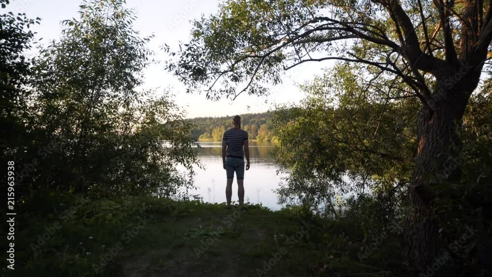 A tall Caucasian male stands by a beautiful lake in Stockholm Sweden. Summer evening, model surrounded by green trees. Steady gimbal footage moving away from the model. Peaceful outdoor area. A tall Caucasian male stands by a beautiful lake in Stockholm Sweden. Summer evening, model surrounded by green trees. Steady gimbal footage moving away from the model. Peaceful outdoor area.