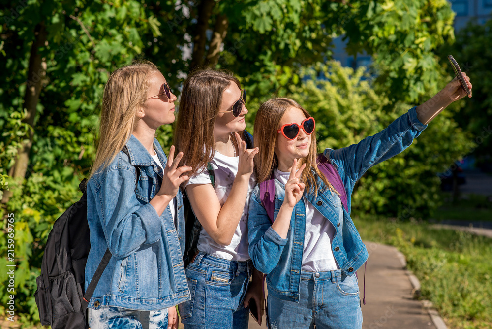 Three girls are teenage girlfriends. In summer after school, in the park. Photographs on a smartphone. Wear jeans clothes and sunglasses. Happy smiling smile they say hello.