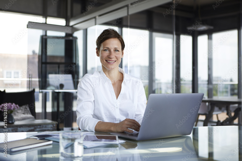 Portrait of smiling businesswoman sitting at glass table in office with laptop Portrait of smiling businesswoman sitting at glass table in office with laptop