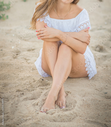 A beautiful model in a white dress is sitting sexually on the sand on a beach