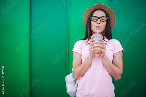 Amazing young woman stands and drinks cold drink. She keeps her eyes closed. Girl is enjoying the moment. She wears hat and glasses. Isolated on striped and pink background. photo