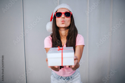 Attractive brunette holds hite box with present in hands. She wears hat and sunglasses. Girl keeps her lips in kiss position. Isolated on striped and white background. photo