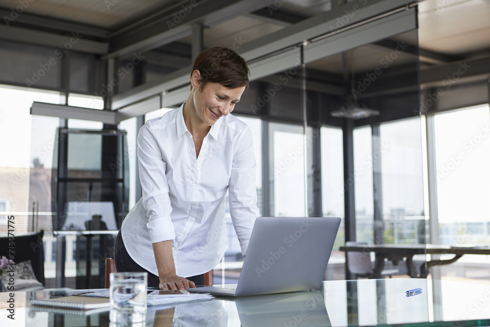 Smiling businesswoman standing at glass table in office looking at laptop Smiling businesswoman standing at glass table in office looking at laptop