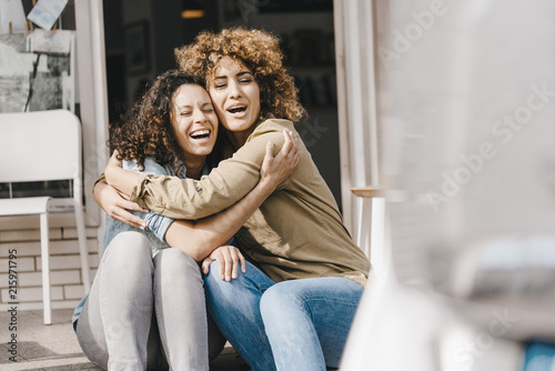 Two laughing friends sitting in front of coworking space, embracing photo