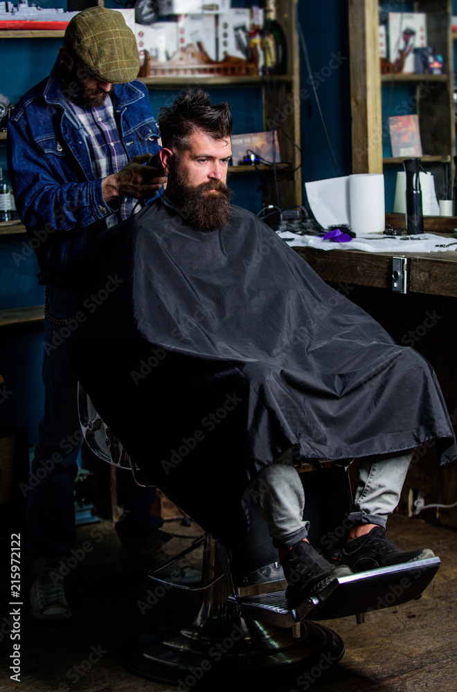 Hipster hairstyle concept. Hipster client getting haircut. Barber with hair clipper works on haircut of bearded guy barbershop background. Barber with clipper trimming hair on nape of client