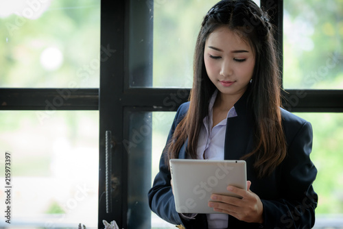 Beautiful Asian businesswoman is using tablet standing near window glass. Within the office. Technology and Business Communication Concept.