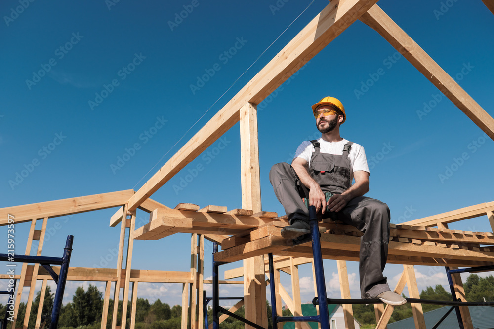 The man builder sits on the edge of the roof of the frame house, in a yellow helmet and gray overalls. The blue sky and clear sunny day.