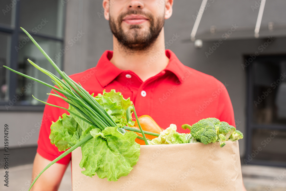 partial view of delivery man in red uniform holding paper bag with fresh vegetables