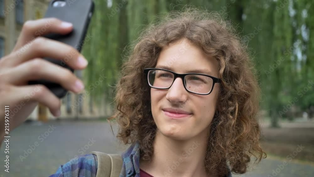 Handsome smart student in glasses with long curly hair taking selfie with his phone, standing on street near university Handsome smart student in glasses with long curly hair taking selfie with his phone, standing on street near university