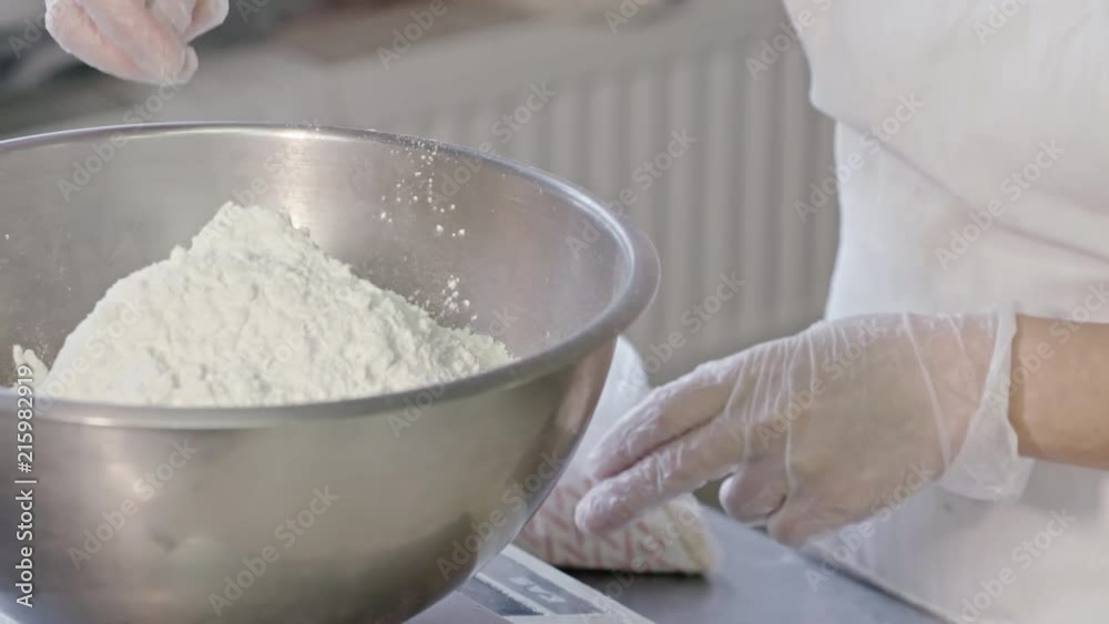 Close-up shot of female baker adding flour into metal bowl while preparing dough Close-up shot of female baker adding flour into metal bowl while preparing dough