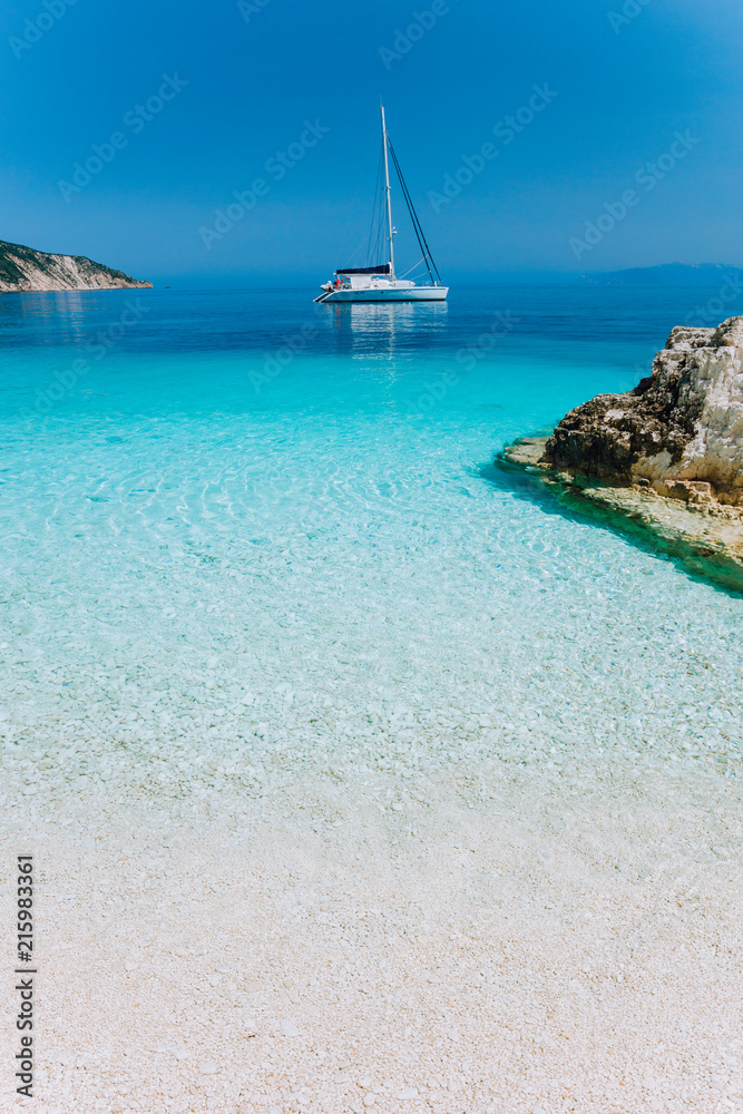 Beautiful azure blue lagoon with sailing catamaran yacht boat at anchor. Pure white pebble beach, some rocks in the sea