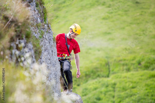 Photo of sporty man looking down in helmet clambering over rock against background of green trees