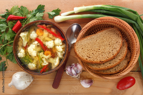 Vegetable soup of cauliflower, carrots, tomato, pepper in a plate with a spoon, bread and onions on a wooden background. Close-up. Top view photo