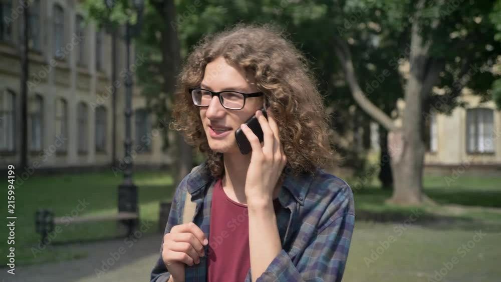 Young happy student with curly hair going to college and talking on phone, walking in park near university, daytime Young happy student with curly hair going to college and talking on phone, walking in park near university, daytime