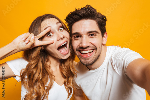 Portrait of attractive man and woman taking selfie photo and showing peace sign, isolated over yellow background
