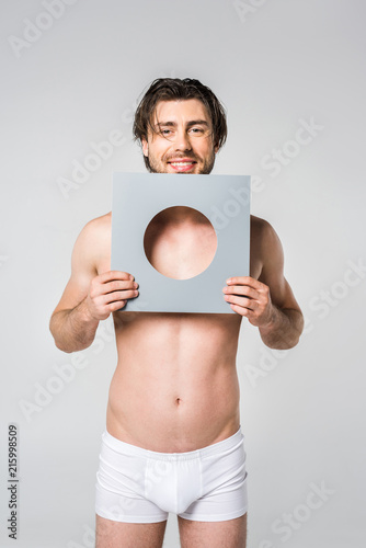 portrait of young smiling man in underwear holding circle paper figure isolated on grey