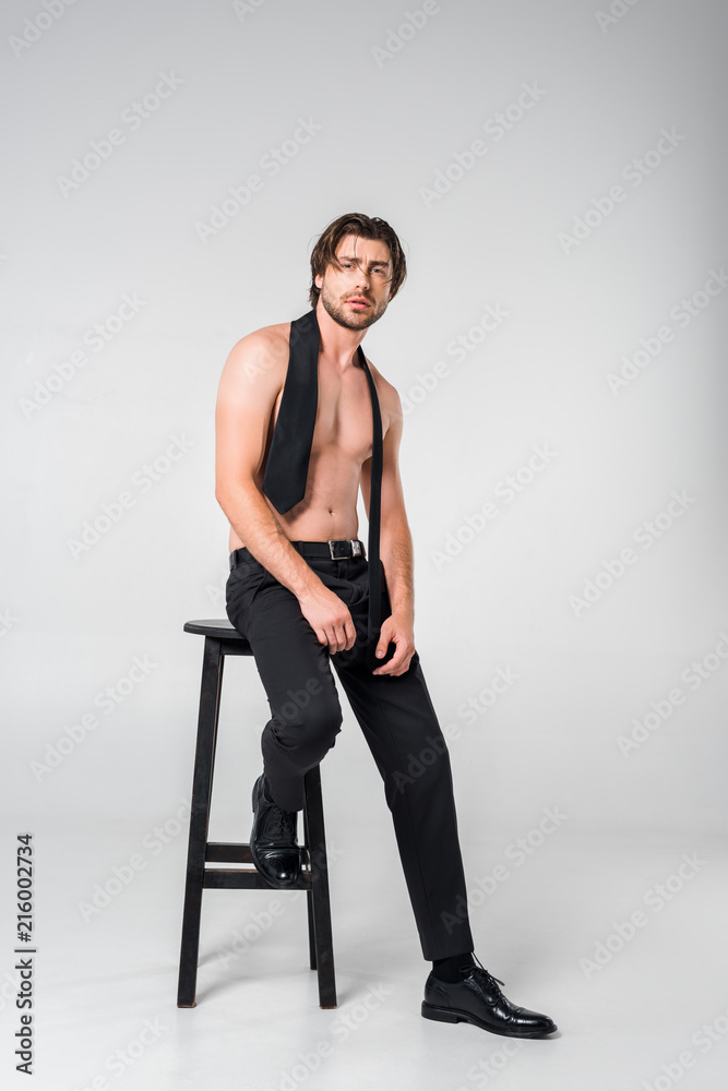 shirtless man in black pants with tie sitting on chair on grey background