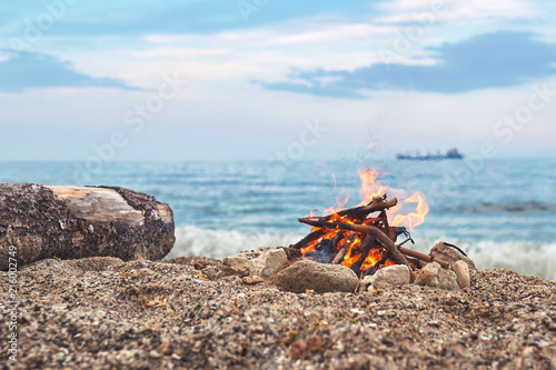 The sea view. A bonfire in the flint of rock stones. A bright fire of a flame in a beautifully built tourist campfire on the sand against the backdrop of a small wave on the sea at sunset