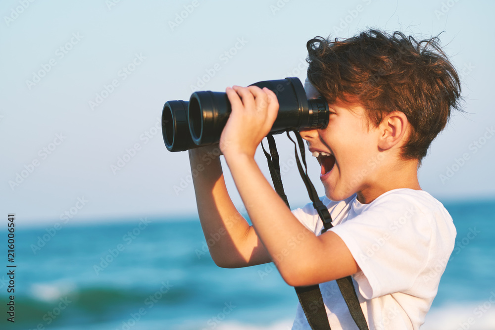 A happy boy holds a large pair of binoculars and stares into the distance on a blue sky against the blue sea and the blue sunset of a sunny day