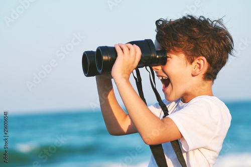 A happy boy holds a large pair of binoculars and stares into the distance on a blue sky against the blue sea and the blue sunset of a sunny day