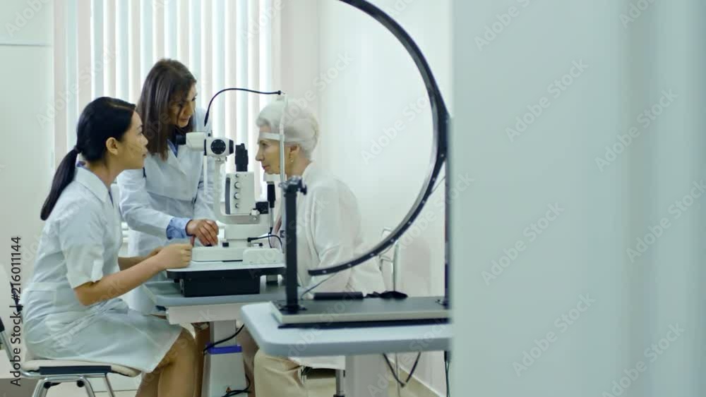 Zoom in of two professional ophthalmologists working together with slit lamp while giving an eye exam to senior woman Zoom in of two professional ophthalmologists working together with slit lamp while giving an eye exam to senior woman