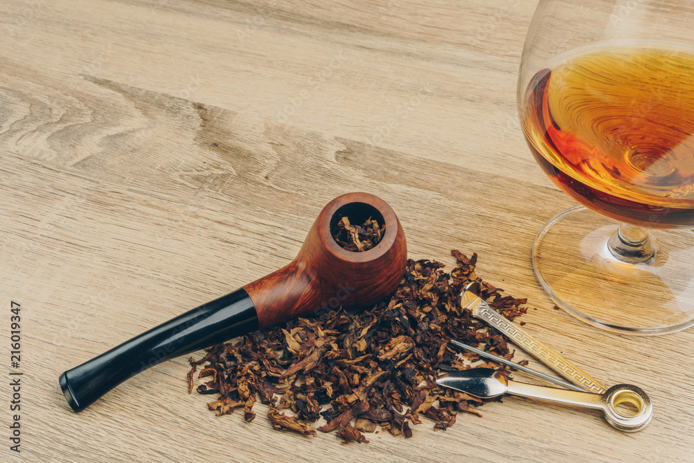 close up of a smoking pipe with tobacco, a pipe tamper tool and a glass of brandy on wooden table