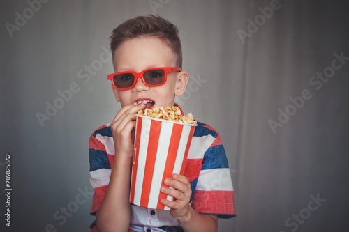 Young boy watch a movie in 3D glasses at the cinema or at home. Little kid eat popcorn over gray background. Home theater. Cute Child in vintage cinema eyeglasses.