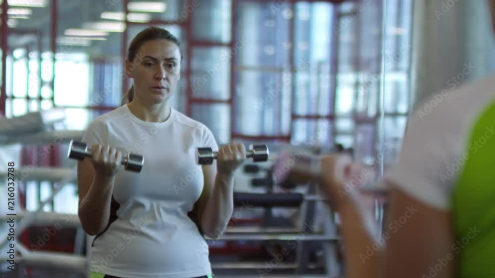 Medium shot with slowmo of middle-aged woman doing dumbbell bicep curls exercise before mirror in gym