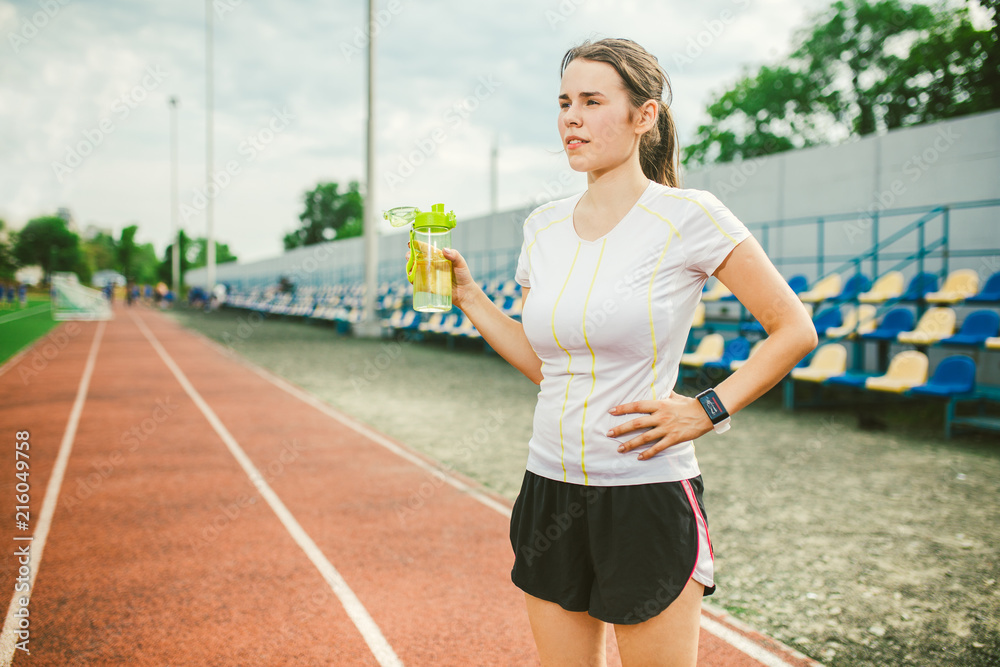 Theme is sport and health. Beautiful young caucasian woman with big breasts  athlete runner stands resting on running stadium, running track with bottle  in hands drinking water in short shorts Stock Photo
