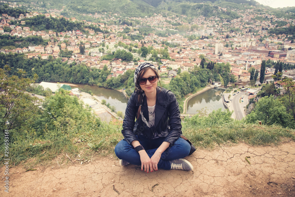 Young casual woman sitting on top of a hill, her back turned to the background of the city Sarajevo in Bosnia