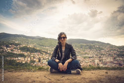 Young casual woman sitting on top of a hill, her back turned to the background of the city Sarajevo in Bosnia