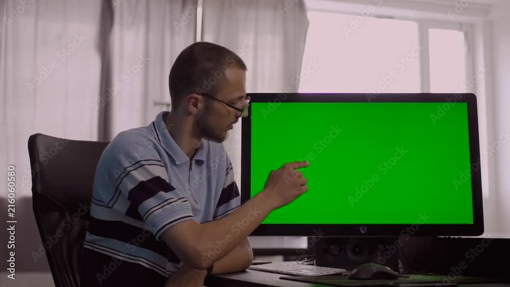 Attractive Caucasian serious young man sitting at computer with big green screen. Nice office worker showing something in monitor, giving thumbs up. Attractive Caucasian serious young man sitting at computer with big green screen. Nice office worker showing something in monitor, giving thumbs up.