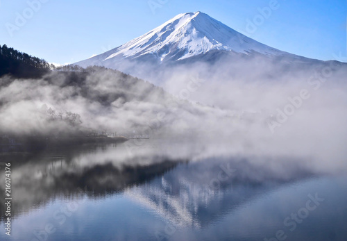 Mount Fuji reflection in calm lake in the early morning. Iconic mountain with snow on top and reflection in calm water. Silhouettes of trees and hills in the mist. Lake Kawaguchiko. Japan.