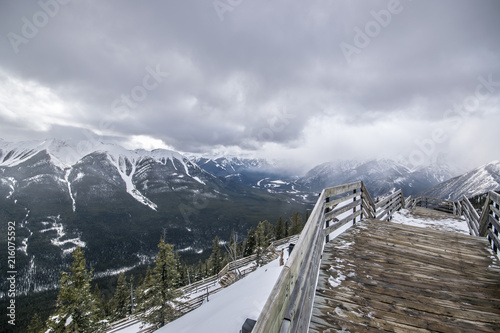 Standing on a walkway looking over the railing at the winter mountain view at Banff National Park. Cloudy skies, snow covered mountain peaks and valley of spruce trees.