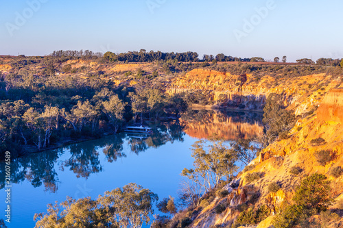 Sandstone eroding cliffs over houseboat moored in Murray River at sunset. Riverland, South Australia