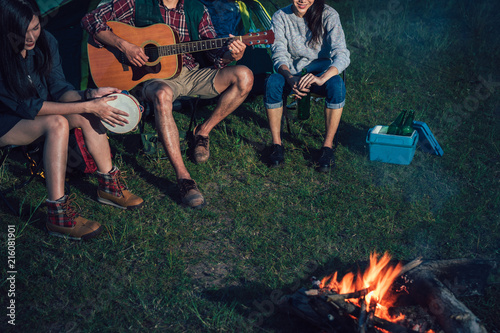 A group of Asian friends sitting on chairs, singing, playing a guitar and drinking some beer and water together outside the tent near the fire while they has camping on Weekend holiday.