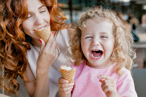 Happy exited little girl with curly hair dressed pink dress laughting and eating ice-cream while sitting with mother in cafeteria