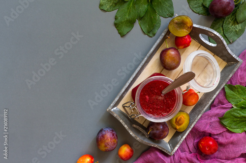 Juicy red plum and plum jam in white utensils and purple runner on gray laconic background pattern with copy space for text flat  lay top view