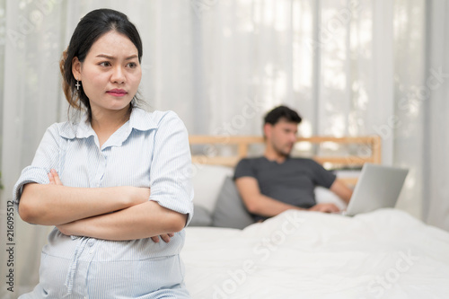 Close Up Of Unhappy Pregnant Woman With Big Belly Over Blur Of A Man Palying Labtop On Bed.Needless Pregnancy And Motherhood Moment People And Expectation. Family planning Concept.In Selective Focus.