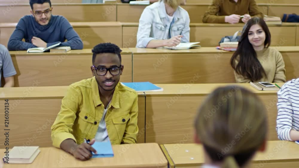 Tilt up shot of multi ethnic group of college students sitting in lecture hall and listening to teacher, one of them asking question