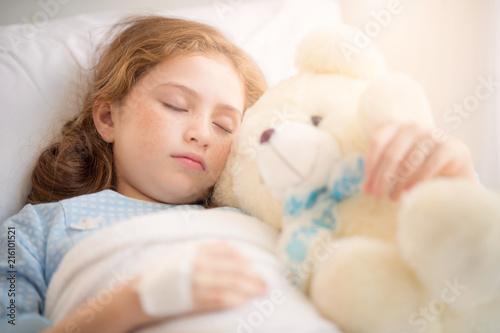 Adorable little girl resting in a hospital bed with her teddy bear. Selective focus at her left eye.