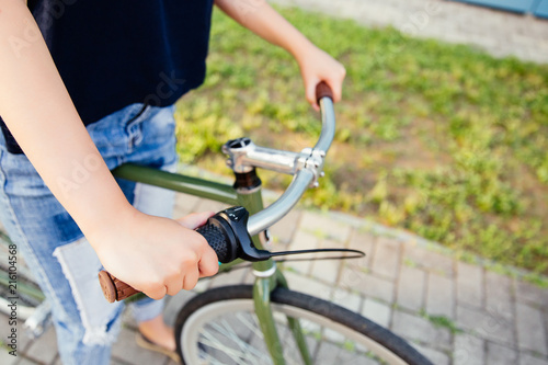 Fototapeta Naklejka Na Ścianę i Meble -  Girl holding the wheel of a bicycle, ready to start riding. Close-up. Dressed in fashionable jeans and t-shirt. Outdoors.