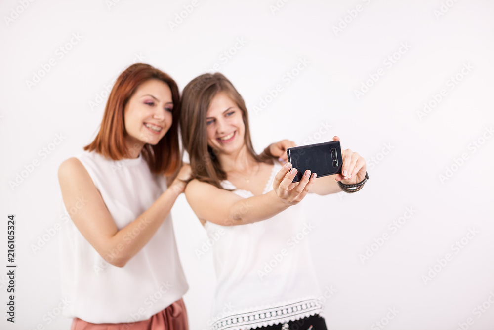 Portrait of two gorgeous young woman, a redhair and a brown-haired, taking a selfie over a white wall in studio Portrait of two gorgeous young woman, a redhair and a brown-haired, taking a selfie over a white wall in studio