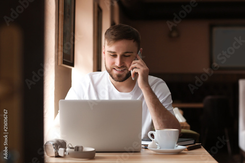 Young freelancer talking on mobile phone while working on laptop in cafe