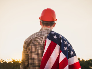 Attractive man in work clothes, holding tools and a US flag in his hands and looking into the distance against the background of trees, blue sky and sunset. View from the back. Labour Day Concept