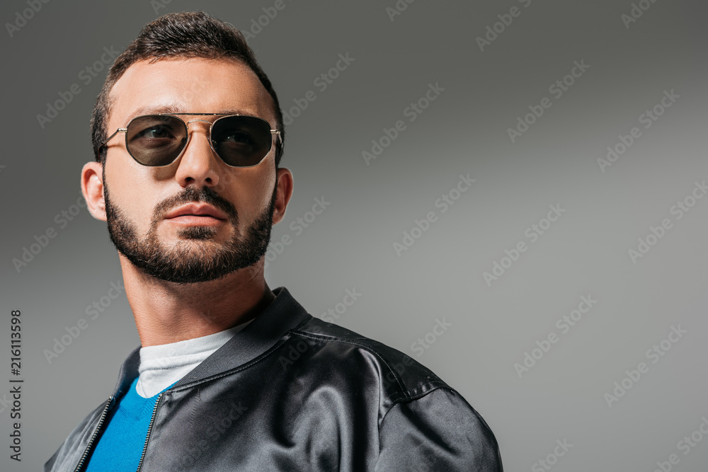 Obraz premium bearded man posing in black bomber and sunglasses, isolated on grey