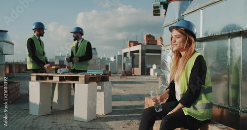 Smiling young woman on construction site , drinking water , wearing a safety helmet and high visibility vest , background builders working photo