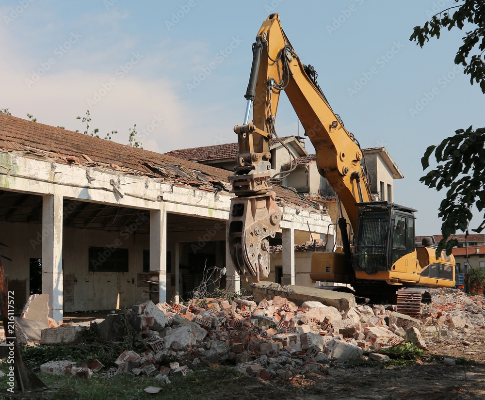 Excavator in front of a semi demolished building, among rubbles. The demolition is done for an urban redevelopment of the area Excavator in front of a semi demolished building, among rubbles. The demolition is done for an urban redevelopment of the area