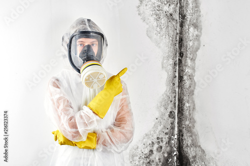A professional disinfector in overalls and a mask points a finger at the black mold on the wall. Removal of the fungus in the apartment and house. Aspergillus..