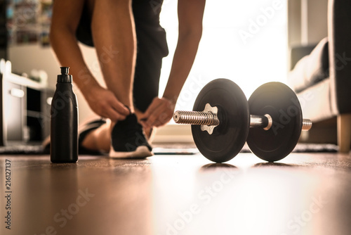 Morning workout routine in home gym. Fitness motivation and muscle training concept. Man in sneakers tying shoelaces in sunlight. Athlete starting exercise with dubbell weight. photo